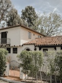 A two-story house with a white stucco facade and a red-tiled roof, surrounded by lush greenery. The architecture features arched windows and a small balcony. A metal gate leads to the entrance, and tall trees provide a natural backdrop.