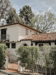 A two-story house with a white stucco facade and a red-tiled roof, surrounded by lush greenery. The architecture features arched windows and a small balcony. A metal gate leads to the entrance, and tall trees provide a natural backdrop.