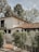 A two-story house with a white stucco facade and a red-tiled roof, surrounded by lush greenery. The architecture features arched windows and a small balcony. A metal gate leads to the entrance, and tall trees provide a natural backdrop.