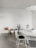 A minimalist room featuring a round white table surrounded by white wire chairs with black cushions. The decor includes several indoor potted plants, with cacti and succulents placed on a stand and the floor. A modern white pendant light hangs from the ceiling, complementing the clean and simple aesthetic.