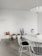 A minimalist room featuring a round white table surrounded by white wire chairs with black cushions. The decor includes several indoor potted plants, with cacti and succulents placed on a stand and the floor. A modern white pendant light hangs from the ceiling, complementing the clean and simple aesthetic.