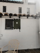 A quaint outdoor patio area featuring a white stucco wall with two small, square windows that have black frames. There is a decorative green window shutter with a diamond cutout next to one of the windows. The roof is lined with brown clay tiles. In the foreground, a beige metal chair with an intricate leaf pattern design is set against the tiled floor.