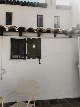 A quaint outdoor patio area featuring a white stucco wall with two small, square windows that have black frames. There is a decorative green window shutter with a diamond cutout next to one of the windows. The roof is lined with brown clay tiles. In the foreground, a beige metal chair with an intricate leaf pattern design is set against the tiled floor.