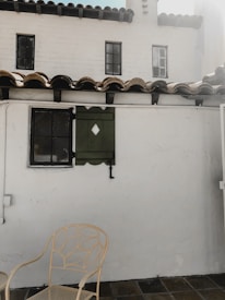 A quaint outdoor patio area featuring a white stucco wall with two small, square windows that have black frames. There is a decorative green window shutter with a diamond cutout next to one of the windows. The roof is lined with brown clay tiles. In the foreground, a beige metal chair with an intricate leaf pattern design is set against the tiled floor.
