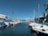 Yachts and boats are docked at a marina flanked by palm trees, with clear blue skies above. A modern building with a geometric facade lines one side of the marina, and a variety of boats are covered with blue tarps.