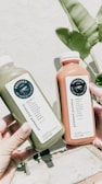 Two hands holding bottles of pressed juice, one green and one orange, against a backdrop of a light-colored wall and green banana leaves.