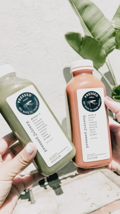 Two hands holding bottles of pressed juice, one green and one orange, against a backdrop of a light-colored wall and green banana leaves.