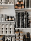 A display of various cylindrical containers and pots arranged on shelves. The items are made from materials like ceramics and metal, featuring textured surfaces and monochromatic color schemes. Some containers have wooden lids, while others are purely decorative.