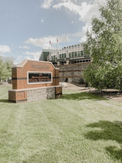 A brick sign with the name 'Appalachian State University' stands on a well-maintained grassy area. Behind it, there is a building labeled 'Holmes Convocation Center' with flags on top, surrounded by greenery and trees. The sky is mostly clear with some fluffy clouds.