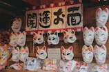 A display of traditional Japanese fox masks, some with autumn leaf designs, arranged on a bamboo background. Above the masks, there is a sign with Japanese characters. The masks are characterized by vibrant expressions and color accents such as red, blue, and yellow.