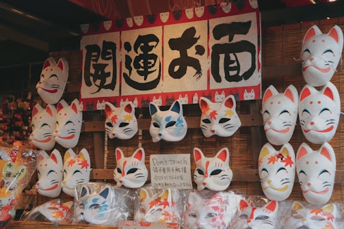 A display of traditional Japanese fox masks, some with autumn leaf designs, arranged on a bamboo background. Above the masks, there is a sign with Japanese characters. The masks are characterized by vibrant expressions and color accents such as red, blue, and yellow.