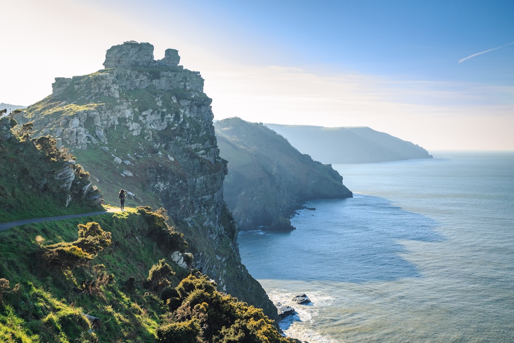 person standing near road viewing cliff and sea under blue and white skies