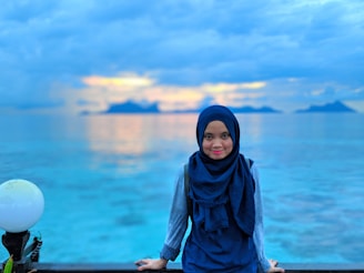 A person wearing a navy blue hijab stands in front of a calm sea with distant islands visible on the horizon. The sky is partly cloudy with hints of a sunset, and the water reflects a soft gradient of blues and oranges. A white spherical lamp is positioned on the left side of the image.