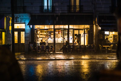 A misty street scene outside the café window, neon signs reflecting on wet pavement under a gentle evening rain.