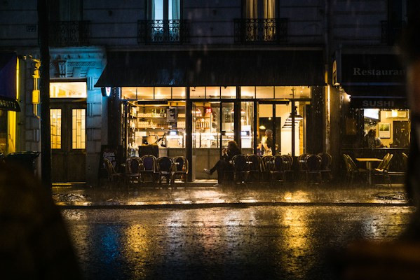 A cozy cafe with warm lighting is visible through large windows, while rain falls heavily outside. Empty chairs line the sidewalk, and a few people are seated inside. The reflections of light on the wet pavement create a serene yet somewhat melancholic atmosphere.