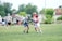 Two lacrosse players are in the foreground engaged in an active play on a grassy field. One player in white and red gear is attempting to maneuver with the ball, while the opposing player in green and navy gear tries to block. Several additional players and spectators are visible in the background, watching the action unfold.