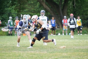 Several athletes are playing lacrosse on a grassy field. Each player is wearing protective gear including helmets and gloves. One player is actively running with a lacrosse stick while opponents attempt to intercept. Other players and spectators are visible in the background, dressed in colorful sports attire.