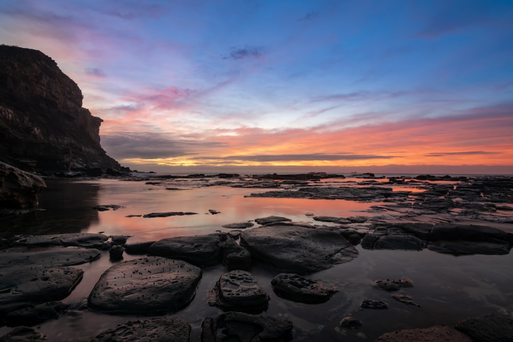 rock formation near sea viewing mountain under orange and gray skies