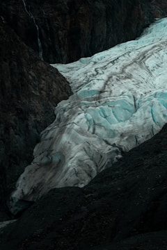 A large glacier with rugged, icy textures flows between dark, rocky cliffs. The ice has a bluish tint, with crevices and ridges visible across its surface, creating a dramatic contrast with the surrounding terrain.