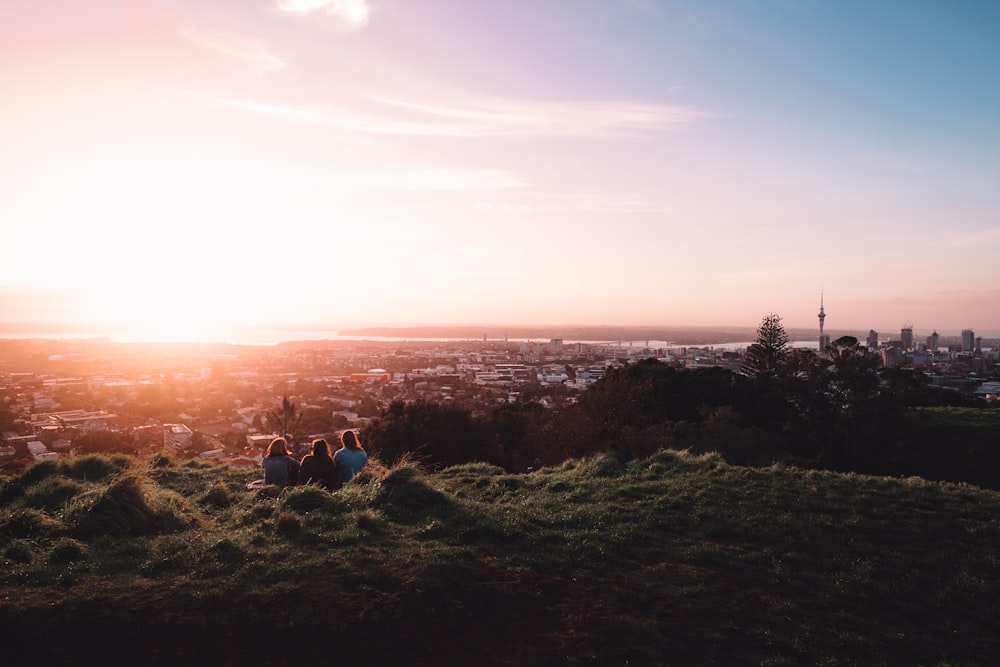 three people sitting on green field facing back on city with high-rise buildings under sunset