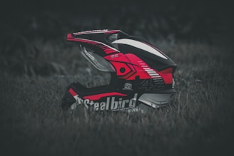 A motocross helmet with vibrant red, black, and white patterns rests on grass. The design is detailed with logos and graphics, and the helmet appears to be well-padded and sturdy.