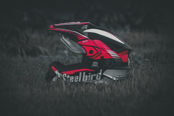 A motocross helmet with vibrant red, black, and white patterns rests on grass. The design is detailed with logos and graphics, and the helmet appears to be well-padded and sturdy.
