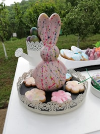 A large decorative bunny figure with a paint splatter design is placed at the center of an ornate tray. Surrounding the bunny are flower-shaped cookies with pastel icing colors. In the background, colorful painted eggs and a small white watering can are visible. The setting appears to be outdoors with lush green foliage.