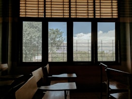 A classroom with empty wooden desks and chairs near a large window. The window has metal grilles and wooden slats at the top, revealing a view of trees and a cloudy sky outside.