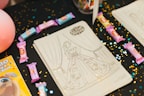 Several coloring sheets featuring a girl in a detailed gown are displayed on a dark tablecloth sprinkled with gold and blue confetti. Various colored candy bars are scattered around the sheets, and there's a partially visible coloring book to the side.