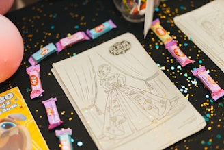 Several coloring sheets featuring a girl in a detailed gown are displayed on a dark tablecloth sprinkled with gold and blue confetti. Various colored candy bars are scattered around the sheets, and there's a partially visible coloring book to the side.