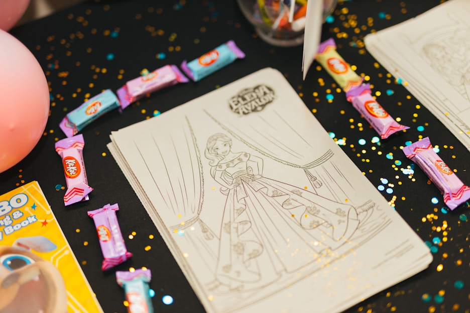 Several coloring sheets featuring a girl in a detailed gown are displayed on a dark tablecloth sprinkled with gold and blue confetti. Various colored candy bars are scattered around the sheets, and there's a partially visible coloring book to the side.