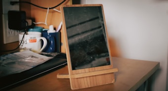 A wooden stand holds a tablet on a desk, with various objects behind it, including mugs, a pen holder, and cables. The background features electrical outlets and a wooden shelf.