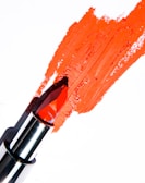 A bright red lipstick is smeared onto a white surface, with its metallic casing visible. The lipstick appears slightly used, creating a textured streak across the background.