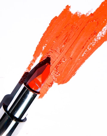 A bright red lipstick is smeared onto a white surface, with its metallic casing visible. The lipstick appears slightly used, creating a textured streak across the background.