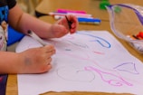 A child is drawing on a white sheet of paper with various colored markers, including black, pink, and blue. The child's hands are holding a black marker while other markers and drawing tools are scattered around on a wooden table.