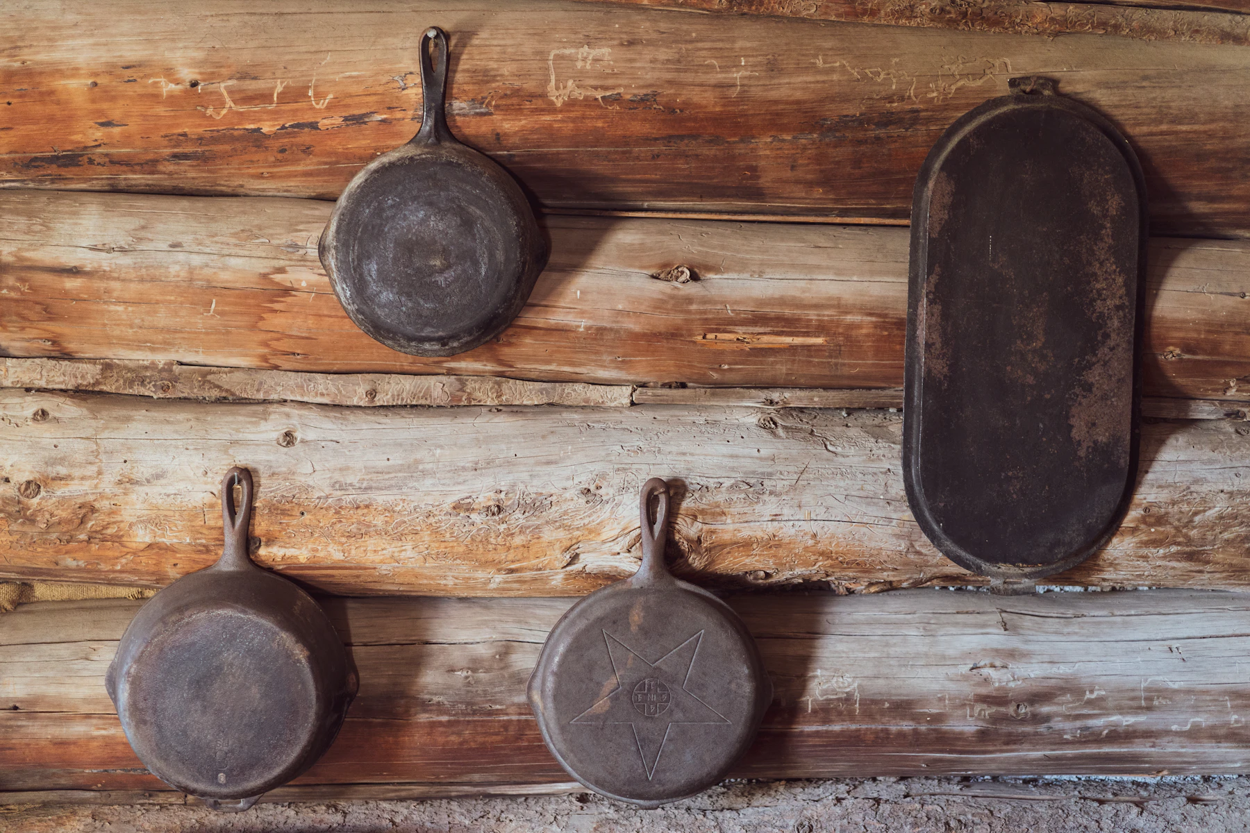 Three old rusty cast iron skillets and an oval griddle hanging on a log-cabin wall, one with a star cast into it