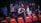 A group of people stands among red and brown chairs labeled 'PASTORES'. Some individuals have their eyes closed and hands placed over their hearts, suggesting a moment of reflection or prayer. The background is dimly lit with focus on the mid-ground figures, who appear to be in an emotional or solemn state. The setting resembles an indoor event or gathering.