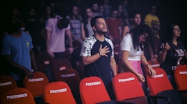 A group of people stands among red and brown chairs labeled 'PASTORES'. Some individuals have their eyes closed and hands placed over their hearts, suggesting a moment of reflection or prayer. The background is dimly lit with focus on the mid-ground figures, who appear to be in an emotional or solemn state. The setting resembles an indoor event or gathering.
