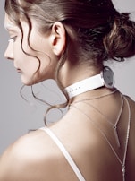 A woman's profile is in focus, showing her hair styled in an elegant bun with loose curls framing her face. She wears a white choker resembling a watch around her neck, and two delicate silver chain necklaces hang down her back. The background is a plain, neutral color, accentuating the minimalist and modern aesthetic.