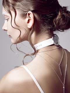 A woman's profile is in focus, showing her hair styled in an elegant bun with loose curls framing her face. She wears a white choker resembling a watch around her neck, and two delicate silver chain necklaces hang down her back. The background is a plain, neutral color, accentuating the minimalist and modern aesthetic.