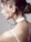 A woman's profile is in focus, showing her hair styled in an elegant bun with loose curls framing her face. She wears a white choker resembling a watch around her neck, and two delicate silver chain necklaces hang down her back. The background is a plain, neutral color, accentuating the minimalist and modern aesthetic.