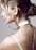 A woman's profile is in focus, showing her hair styled in an elegant bun with loose curls framing her face. She wears a white choker resembling a watch around her neck, and two delicate silver chain necklaces hang down her back. The background is a plain, neutral color, accentuating the minimalist and modern aesthetic.