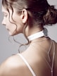 A woman's profile is in focus, showing her hair styled in an elegant bun with loose curls framing her face. She wears a white choker resembling a watch around her neck, and two delicate silver chain necklaces hang down her back. The background is a plain, neutral color, accentuating the minimalist and modern aesthetic.