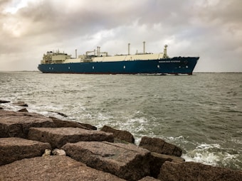 A large cargo ship with a dark blue hull and white superstructure is sailing on a calm sea. The sky is overcast with grey clouds, indicating possible stormy weather. In the foreground, large rocks are visible near the shoreline where waves are gently crashing.
