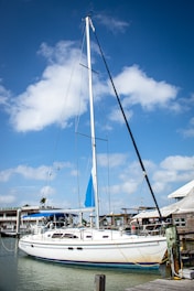 A sailboat is docked at a marina under a clear blue sky with scattered white clouds. The boat has a tall mast and a blue sail, and is surrounded by wooden docks and small buildings.