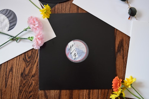 A wooden surface is adorned with vinyl records and colorful flowers. One black vinyl record features prominently at the center, surrounded by white vinyl sleeves. Brightly colored flowers, including pink, yellow, and orange blooms, add a touch of vibrancy. A pair of round sunglasses rests casually on the open white sleeve.