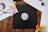 A wooden surface is adorned with vinyl records and colorful flowers. One black vinyl record features prominently at the center, surrounded by white vinyl sleeves. Brightly colored flowers, including pink, yellow, and orange blooms, add a touch of vibrancy. A pair of round sunglasses rests casually on the open white sleeve.