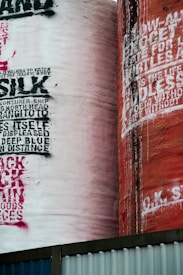 Two large industrial silos feature prominently, with one painted in a light pink shade and the other in a rusty orange. Both are covered in bold, expressive graffiti with a variety of font styles and sizes. The silos stand against a backdrop of blue and white metal barriers.