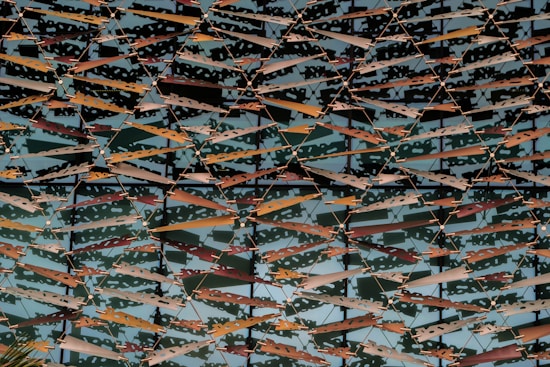 An intricate pattern of metal panels is arranged in a geometric, lattice-like design. The panels are a mix of warm colors, including reds, oranges, and browns, alongside neutral hues. They overlap in layers, creating a dynamic and abstract visual of light and shadow play. The structure is likely part of an architectural facade.