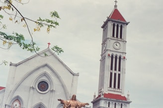 The image presents a church building with a prominent clock tower featuring red-tiled roofs and a cross on top. The facade includes an arched window with stained glass. In the foreground, a statue of Jesus with arms outstretched is visible, and branches with green leaves frame the upper left corner.